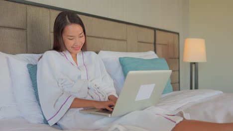 Una-Joven-Asiática-Sentada-En-La-Cama-De-Su-Hotel-Y-Usando-Una-Bata-Equilibra-Su-Laptop-En-Sus-Piernas-Mientras-Toca-El-Teclado