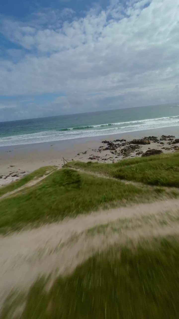 FPV drone Brittany beach house sits in lush gardens with pine trees, facing sandy beach and calm Atlantic waves under blue sky, peaceful coastal living in France