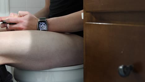 young white male sitting on a toilet addicted to his cell phone and smartwatch