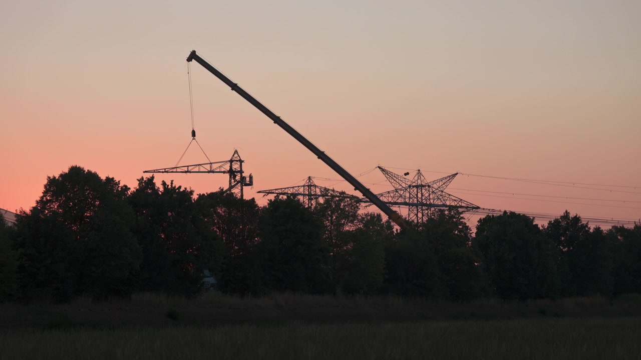 Industrial climbers are working on a power pole with an angle grinder. One arm of the pole is suspended from a truck-mounted crane and is being dismantled as the sun sets.