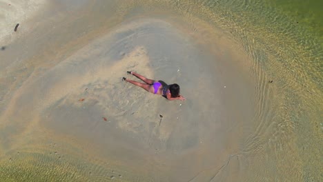 Vista-Aérea-De-Una-Mujer-Joven-Tendida-En-Un-Banco-De-Arena-En-Un-Río-Usando-Solo-Un-Sexy-Bikini-Morado