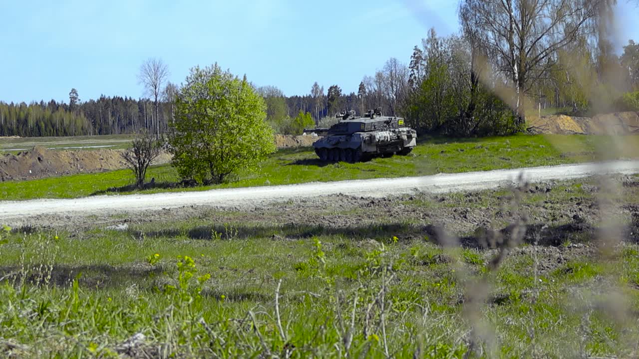 Gorgeous footage of a British army military Challenger 2 4034 tank hidden behind some bushes in a grassy green field and aiming at a tree line in the back with its large cannon during sunny warm day.
