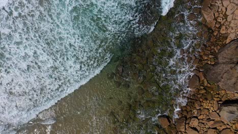 aerial-drone-flight-on-a-sunny-summer-day-over-a-cliff-along-which-the-beach-of-orange-stones-runs-against-which-the-waves-of-white-foam-and-turquoise-sea-calmly-collide-in-Cantabria-Spain