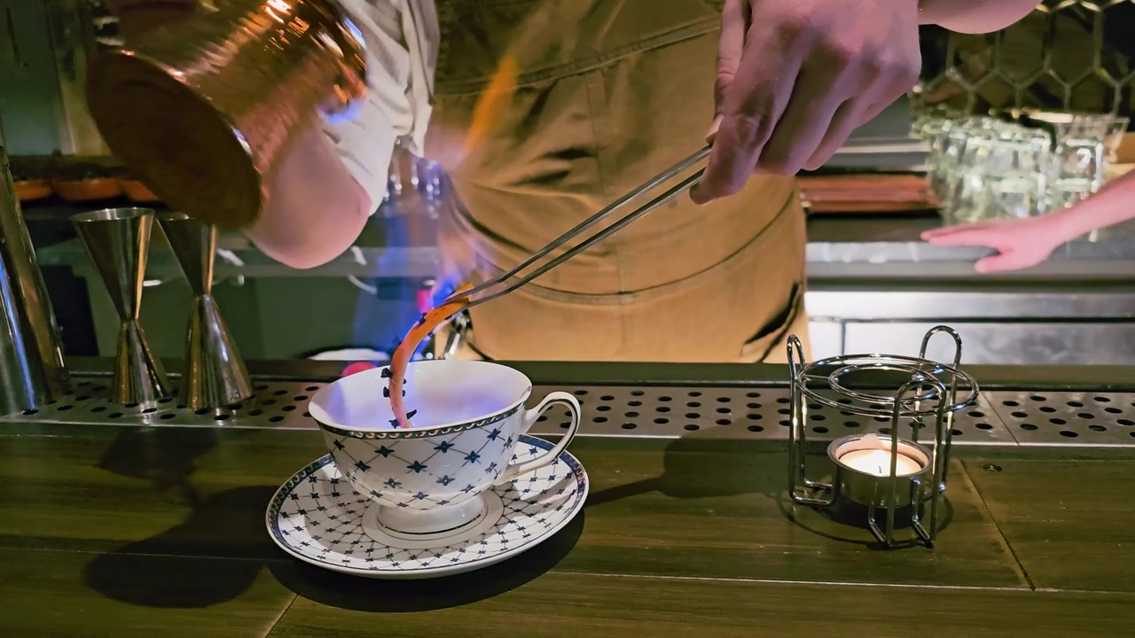 Un barman verse un liquide enflammé d'un pot en cuivre dans une tasse à thé à motifs, avec une flamme bleue, un chauffe-bougie, des pinces métalliques, un éclairage de bar chaleureux et un service de cocktail au thé spectaculaire.