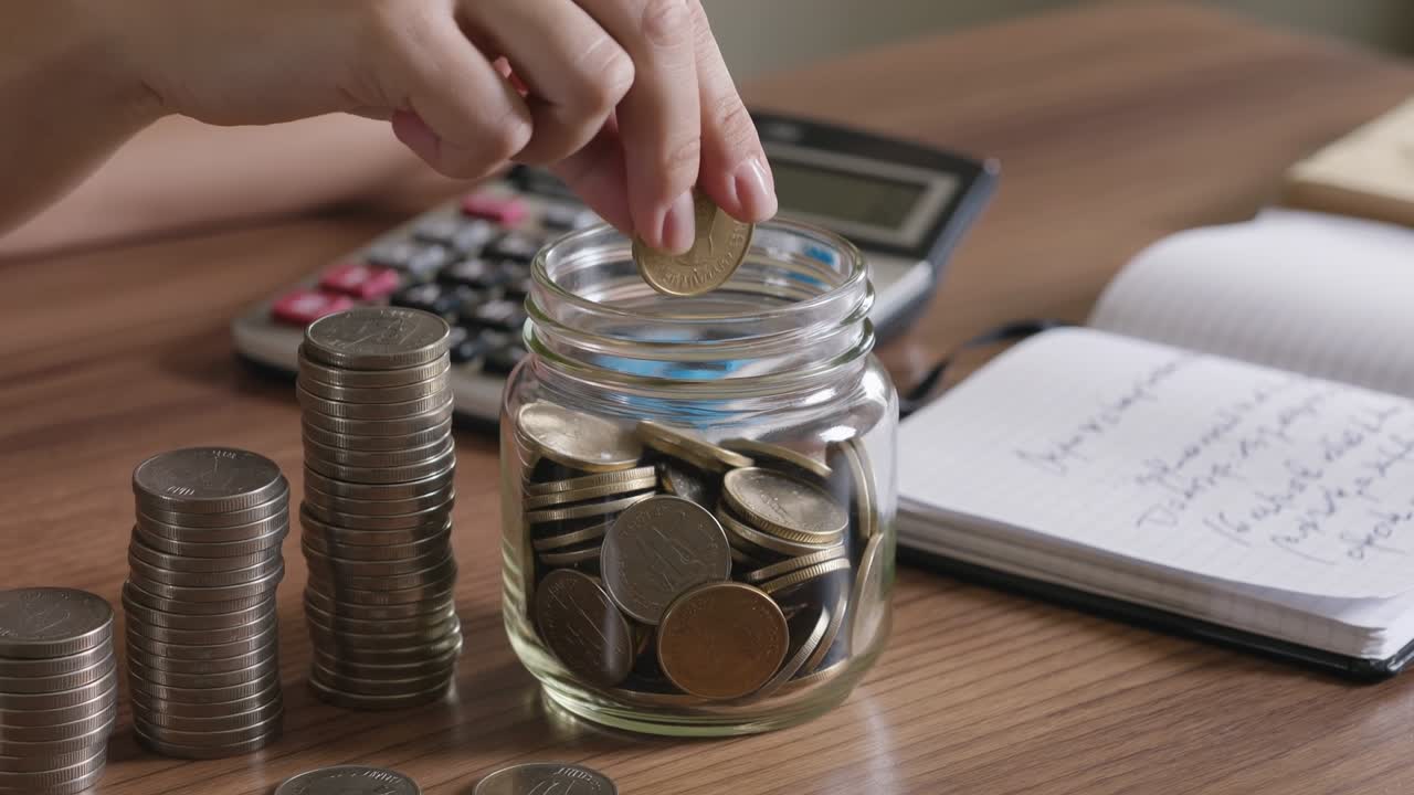 Person putting coins into a glass jar for savings with a calculator and money stacks on desk