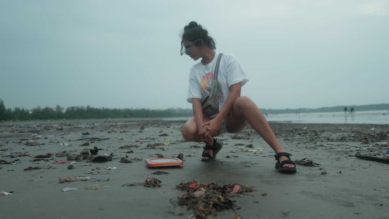 Ragazza coscienziosa accovacciata che guarda i rifiuti sulla spiaggia di sabbia nera in una giornata nuvolosa
