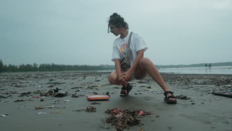 Chica-Concienzuda-En-Cuclillas-Mirando-La-Basura-En-La-Playa-De-Arena-Negra-En-Un-Día-Nublado