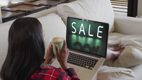 biracial woman on couch with coffee, using laptop for online shopping, slow motion