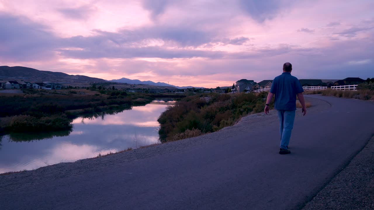 hombre maduro caminando por un sendero al lado de un río durante una increíble puesta de sol - lejos de la cámara estática