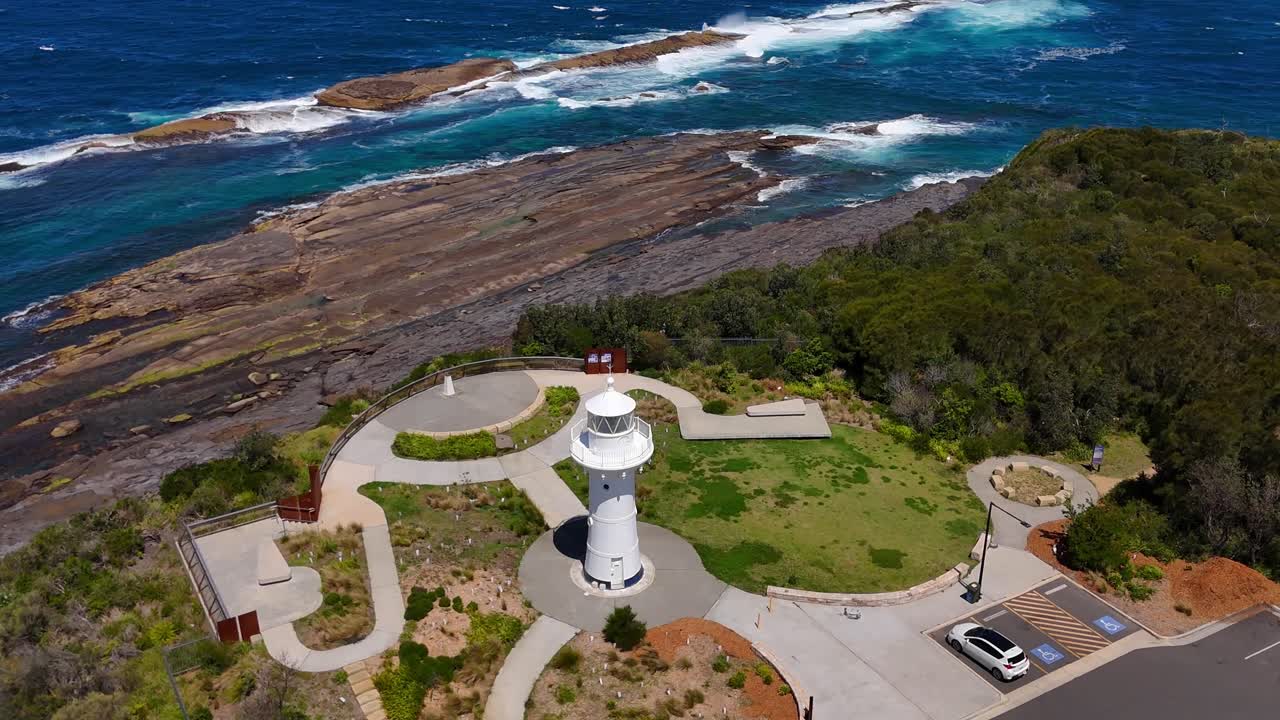 Lighthouse stands above rugged point with swirling surf and coastal scrub surrounding the headland, aerial orbit of Warden Head Lighthouse