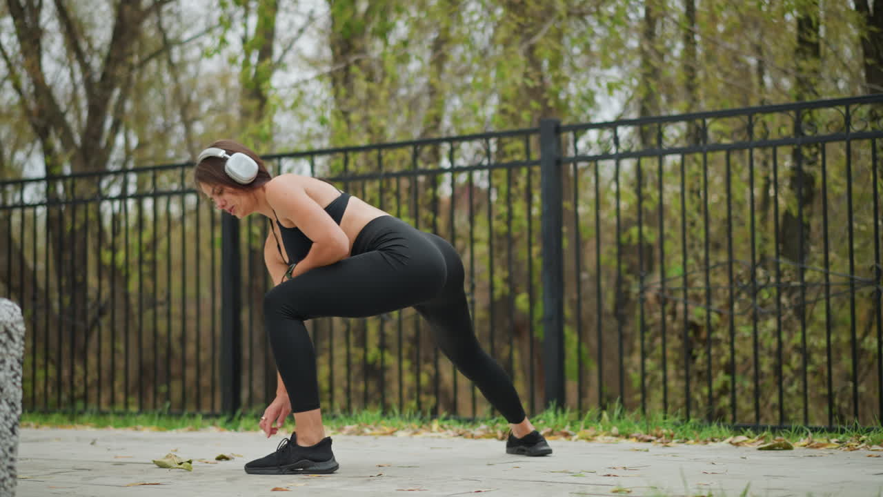 Young girl in black fitness wear stretching and listening to music with headphones, outdoor workout in park, iron fence and grass visible in background, exercising for fitness and health