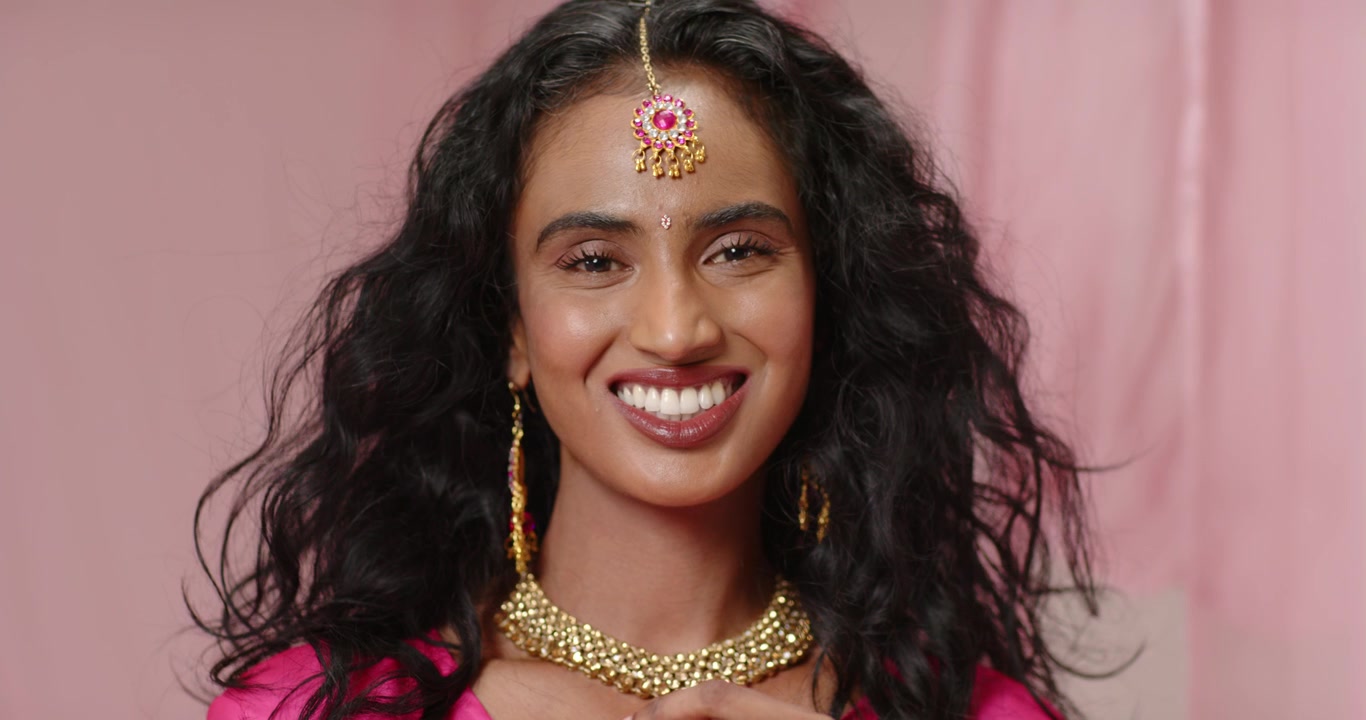 Indian woman posing in studio in pink dress tilting head touching necklace, smiling showing jewelry
