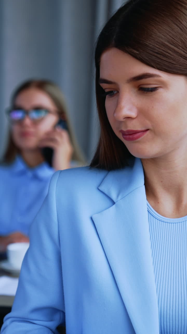 Lady office employee puts off her paper notebook and takes laptop. Working hours in office. Female colleague speaks on the phone at backdrop in blur. Vertical video