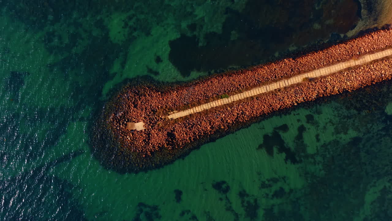 Dæmning af sten og belagt sti for besøgende. Droneflyvning over molen. Havgrønt vand omkring.