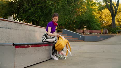 una chica joven con un corte de pelo corto en una parte superior púrpura saca patines rosados de una mochila amarilla y los pone en un área especial para patinadores en el parque en verano