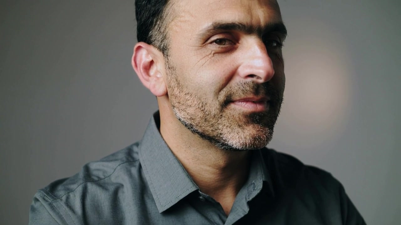 Uomo che porta le mani al colletto e sistema la camicia grigia per comodità, chiudendo gli occhi in studio, con luce soffusa.