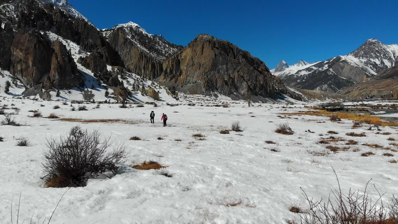Two travelers trekkers walking hiking through a snow field. Sunny day in winter. Annapurna Conservation Area, Manang Valley, Nepal. Aerial Drone shot closing in.