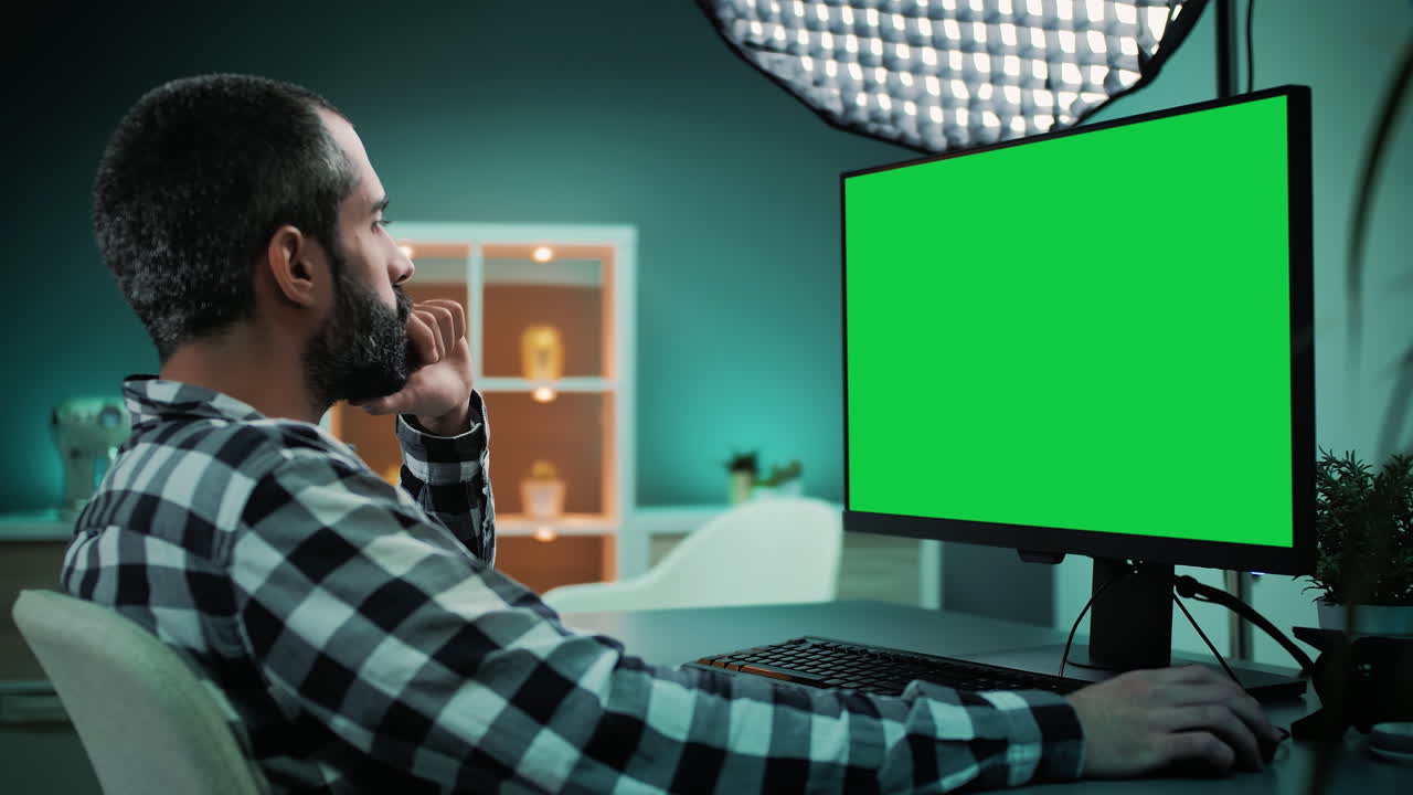 Slow motion shot over the shoulder of a handsome and focused young freelancer, copywriter, designer, editor or content creator working on desktop computer with chroma key green screen mock up display