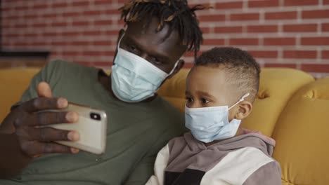 father and son are sitting on a sofa in medical face masks, watching video on smartphone, close up
