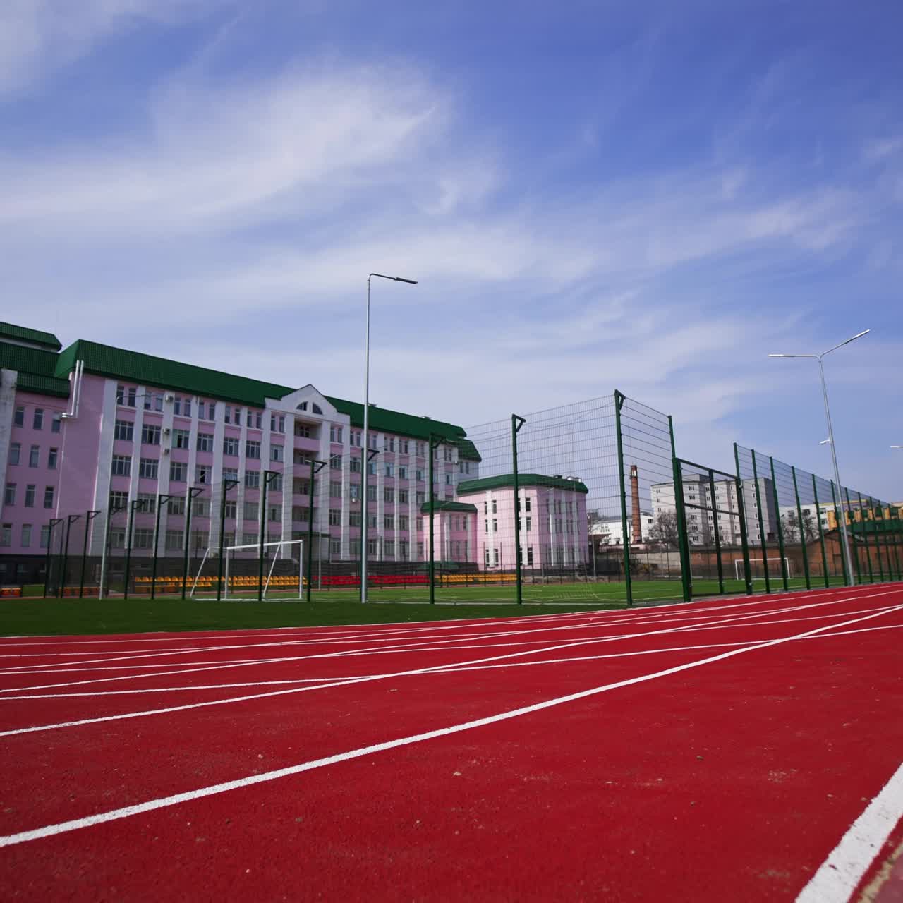 Approaching a soccer field surrounded by fence in front of the big pink building. Red running track at the foreground. Low angle view