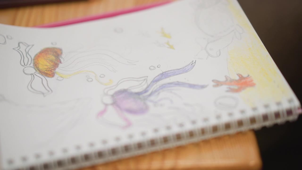 Close-up of tilted spiral-bound sketchbook page featuring ink and colored pencil drawings of whimsical creatures with visible pencil outlines, soft shading, eraser marks, warm light, and subtle page