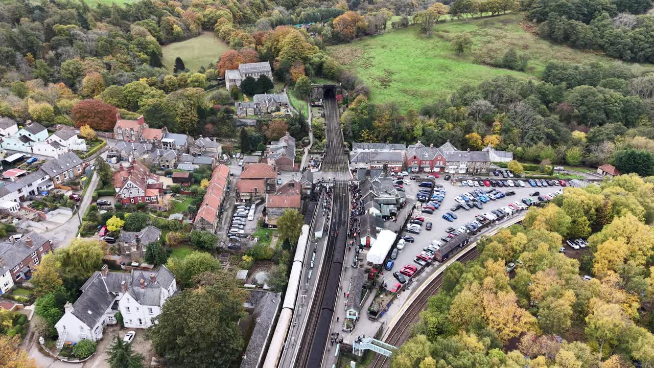 Aerial drone video showing Pickering heritage railway station in North Yorkshire, England, during autumn, featuring historic trains, village buildings, parking area, and colorful countryside landscape
