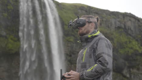 man controls an fpv drone for aerial photography and video with a remote control and goggles, behind a waterfall in iceland