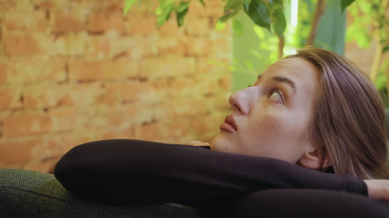 Young lady with relaxed posture leans on cushion and gazes upward in cozy green space filled with soft natural light, brick wall , and hanging indoor plants evoking calm and thoughtful ambiance