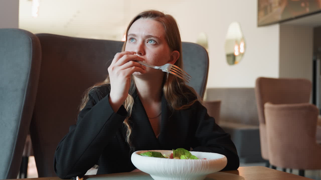 Donna d'affari che gusta un'insalata in un caffè, donna composta in blazer seduta in poltrona, ciotola bianca di verdure, sguardo pensieroso tra un boccone e l'altro, interno luminoso di un bistrò, luce naturale, atmosfera raffinata e rilassata