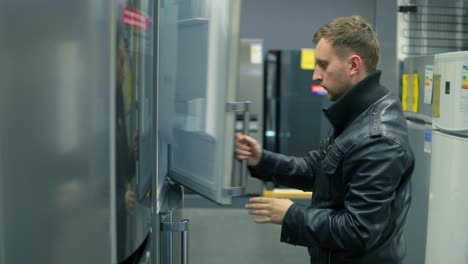 Young-man-is-choosing-a-refrigerator-in-a-store.-He-is-opening-the-doors,-looking-inside