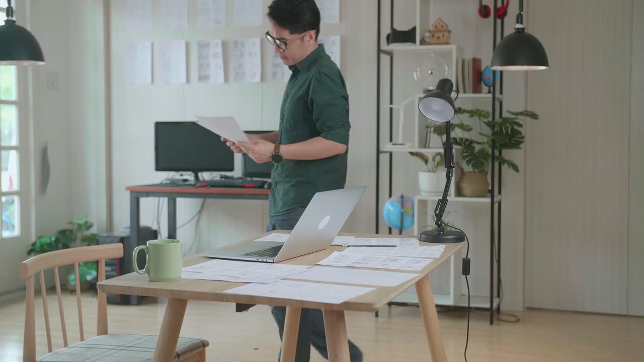 Creative Asian Designer Works On A Storyboard, Looks At His Sketches And Concepts, Choosing The Best Drawings For His Project. Video Editing, Comics Compilation, Application Ui Or Game Plot