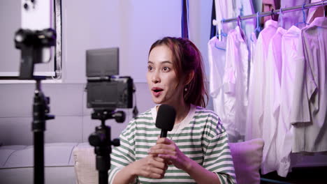 a live streaming girl promoting her merchandise. young asian lady using mobile phone receiving purchase order and showing clothes while live. small business owner with online market delivery concept.