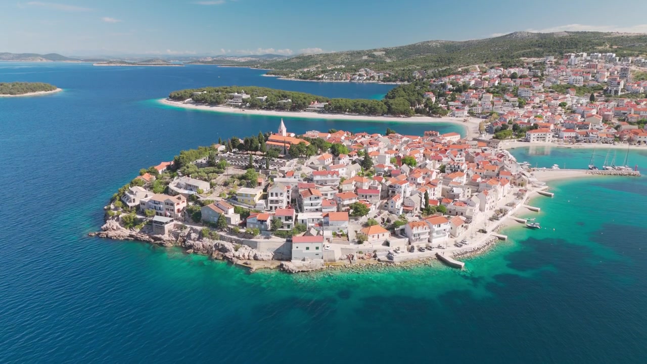 Dron nad Jadranem s výhledem na poloostrov starého města Primošten, zvedá se, aby odhalil kostelní věž a úzké pozemní spojení v jasném pobřežním světle.