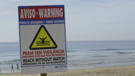 Advertencia-De-Peligro-De-Ahogamiento-En-Portugués-E-Inglés-En-La-Playa-Fonte-Da-Telha-En-Portugal-Con-Ciclista,-Pareja-Y-Perro-En-La-Distancia