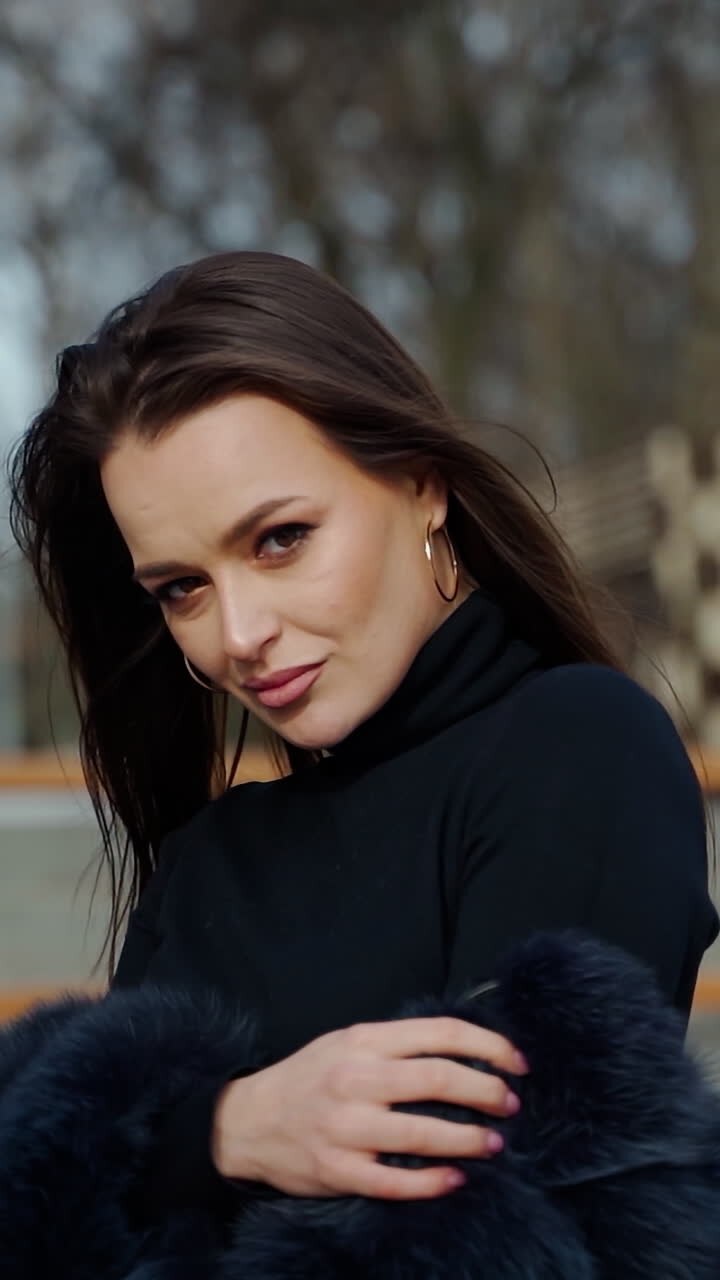 Portrait of beautiful model. Glamour girl in black fur coat posing on camera outdoors. Attractive brunette in the park. Camera moves around. Vertical video