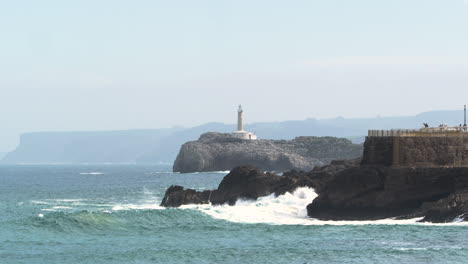 white lighthouse in distance while ocean waves hit rocky spanish coastline