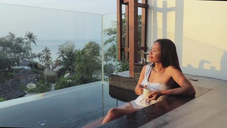 young girl enjoying a bath on a balcony in a luxurious hotel, holding a cup of coffee or tea and admiring a tropical view