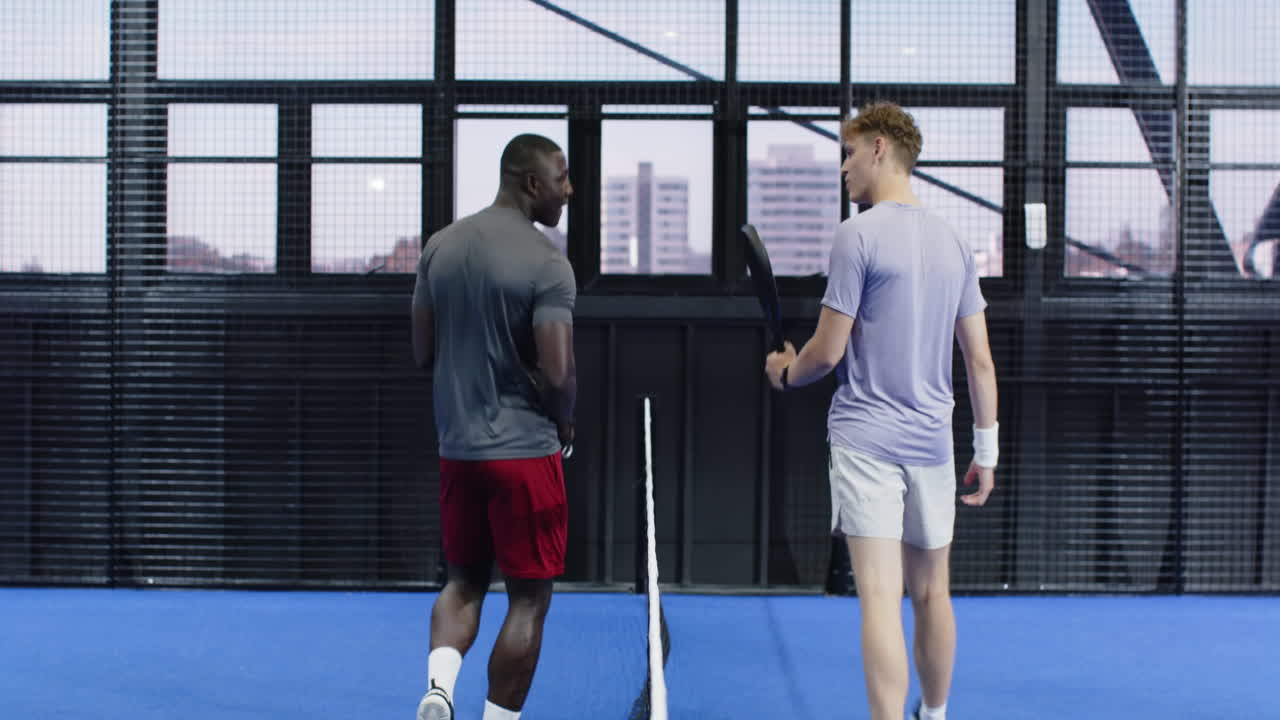 Two diverse men playing padel tennis on indoor court, enjoying friendly conversation on court