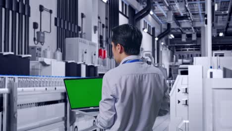 back view of an asian male professional worker standing with water production in bottling factory, typing on laptop's keyboard with green screen and look at the factory once