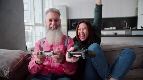 retrato de un hombre mayor feliz con cabello gris y una barba exuberante en una camisa rosa que juega videojuegos usando joysticks con su hija adulta morena en una chaqueta verde oscuro en un sofá marrón en un apartamento moderno