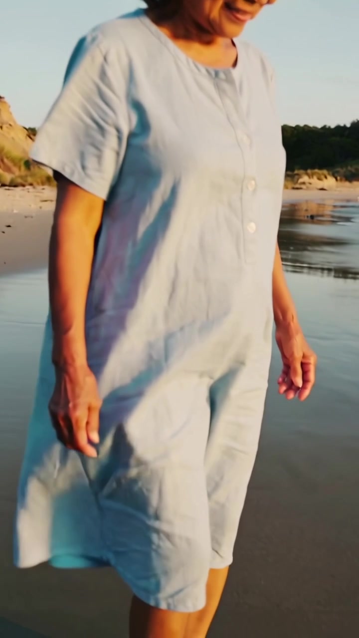 Video verticale: Donna asiatica anziana che cammina, guidata dal sole caldo verso le onde, con un vestito azzurro.