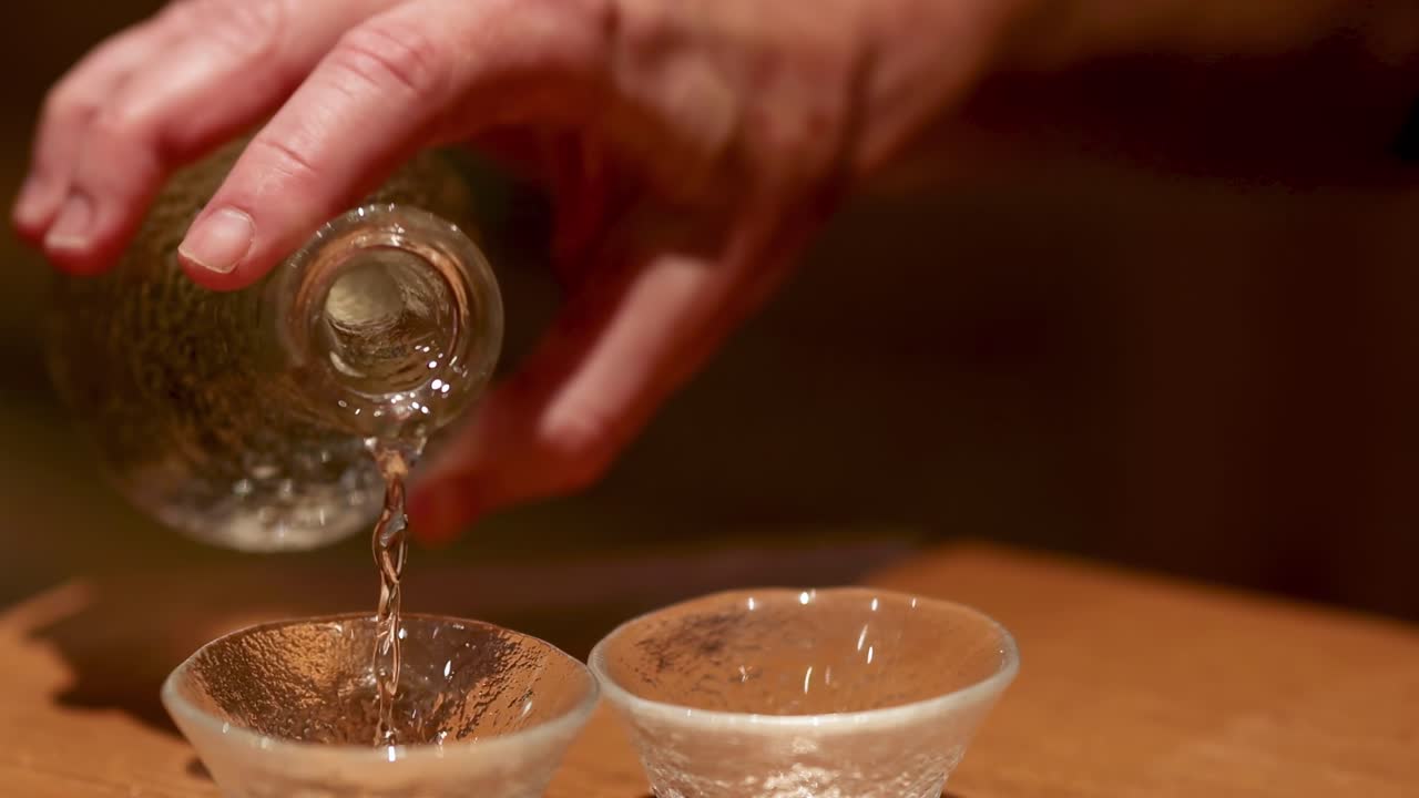 A sequence showing a hand pouring clear liquid from a textured bottle into two small glass cups.