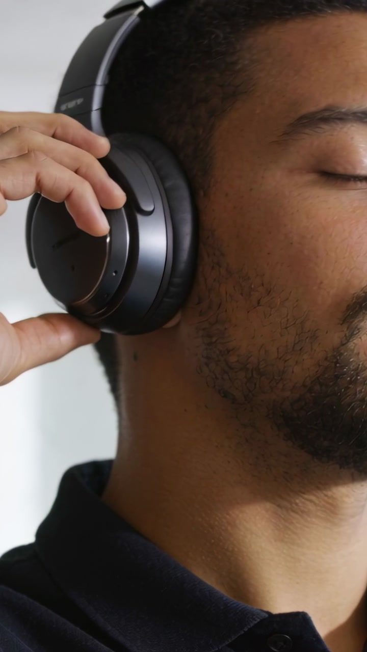 Vertical video: Touching control area, man adjusting headphones at home for comfort, in dark polo
