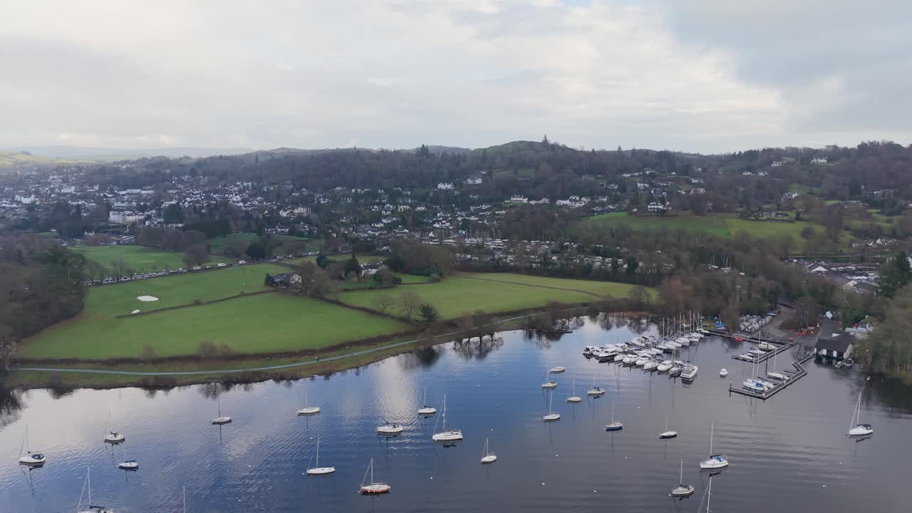 Drone terbang menghadap ke depan di atas Danau Windermere, di atas yacht pada tambatan ayun menuju tepi pantai Bowness. Pepohonan musim dingin, ladang, rumah, dan perbukitan yang jauh berada di bawah langit abu-abu dan putih yang berawan