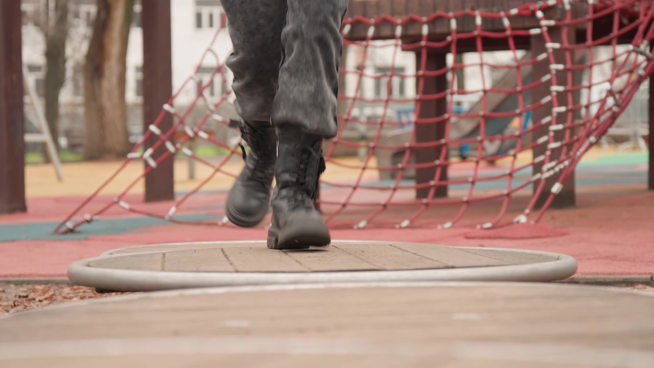 Child stepping toward circular platform by rope net, playground scene shows cautious approach, boots on deck, rope climbing structure and soft rubber surface under pale winter sky, atmosphere