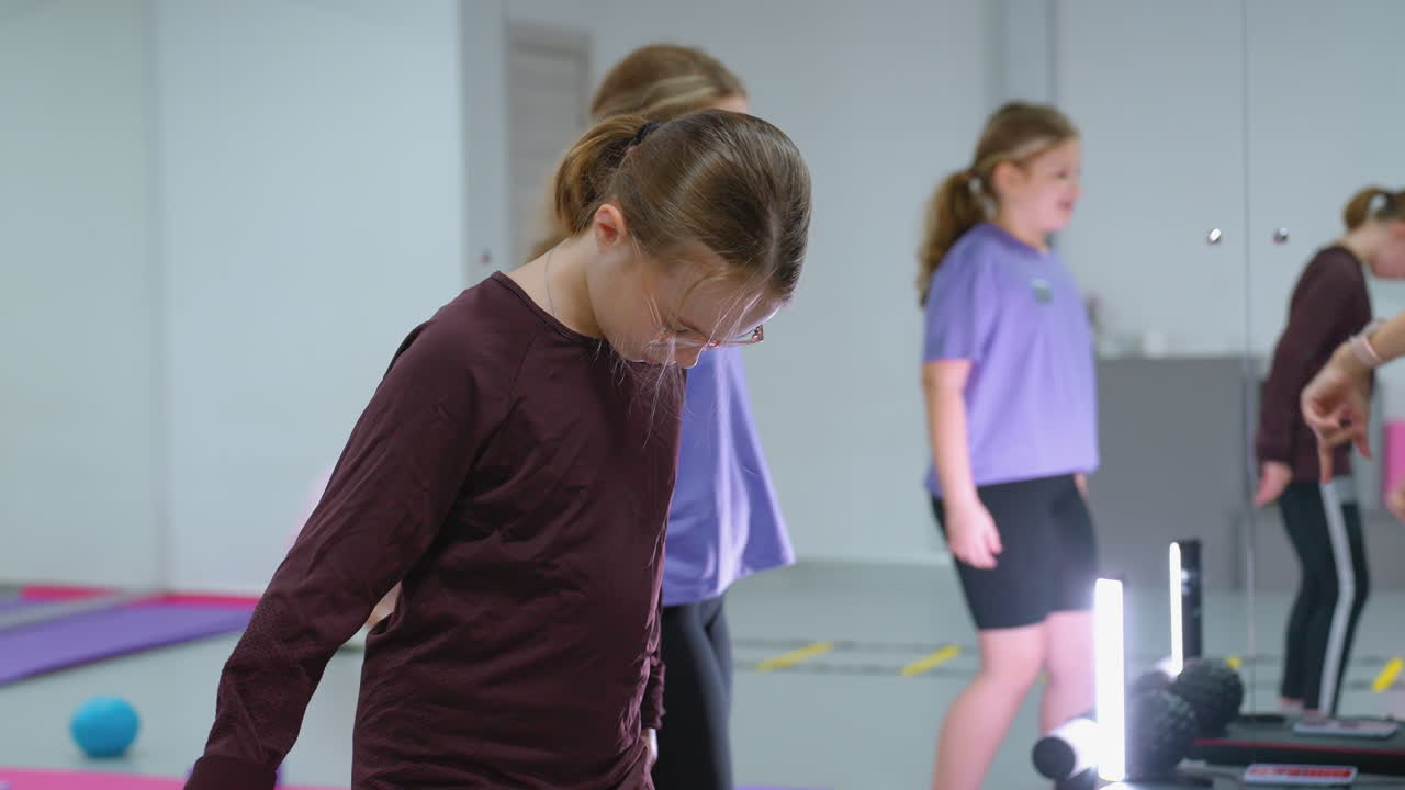 instructor con reloj inteligente guía a los niños en la clase de fitness centrarse en la niña de púrpura pisando el bloque mientras otro niño mira atentamente, el reflejo visto a través del vidrio