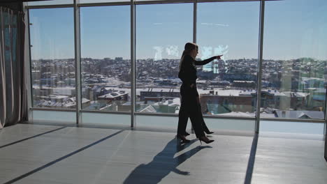 Two ladies coworkers go along the panoramic windows. Female colleagues communicate in the spacious office. Cityscape at the backdrop.