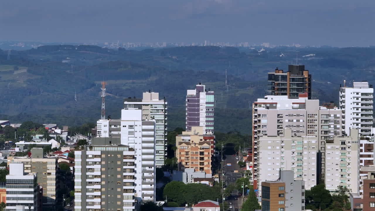 Veduta aerea con teleobiettivo dello skyline di Bento Gonçalves con torri residenziali, colline stratificate e Garibaldi visibile all'orizzonte lontano.