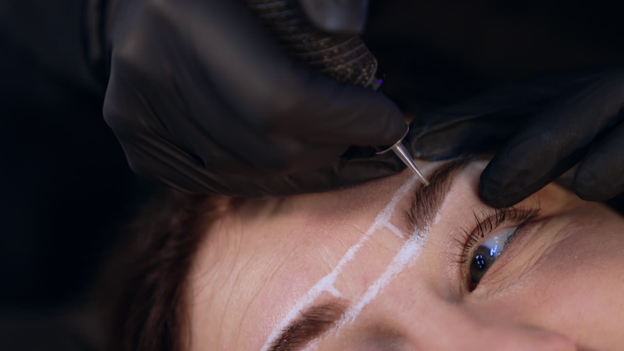 Cosmetologist uses special tool to perform permanent eyebrow make up procedure. Client's eyebrows are lined with white pen. Close up.