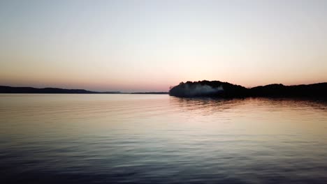 Un-Lago-Tranquilo-Al-Atardecer,-Que-Refleja-Los-Suaves-Colores-Pastel-Del-Cielo-Con-La-Silueta-De-Una-Pequeña-Isla-En-La-Distancia.